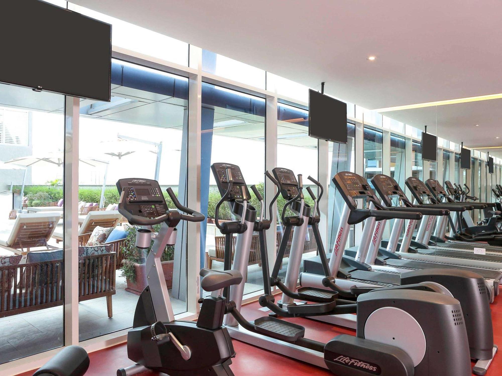 Novotel Jazan Exterior foto The photo shows a modern gym interior with several exercise machines, specifically elliptical trainers, arranged neatly in rows. Large windows allow natural light to fill the space, providing a view outside to a patio area with lounge chairs. There a