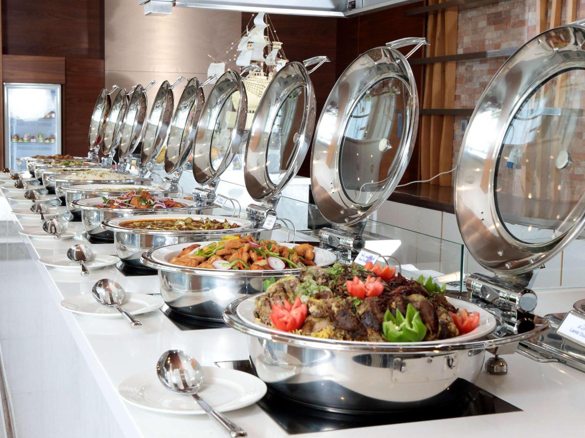 Novotel Jazan Exterior foto The photo shows a buffet setup with a long row of stainless steel chafing dishes. Each dish has a lid, and they are arranged neatly on a white countertop. The food inside appears to be a variety of dishes, including a colorful mix of vegetables and p