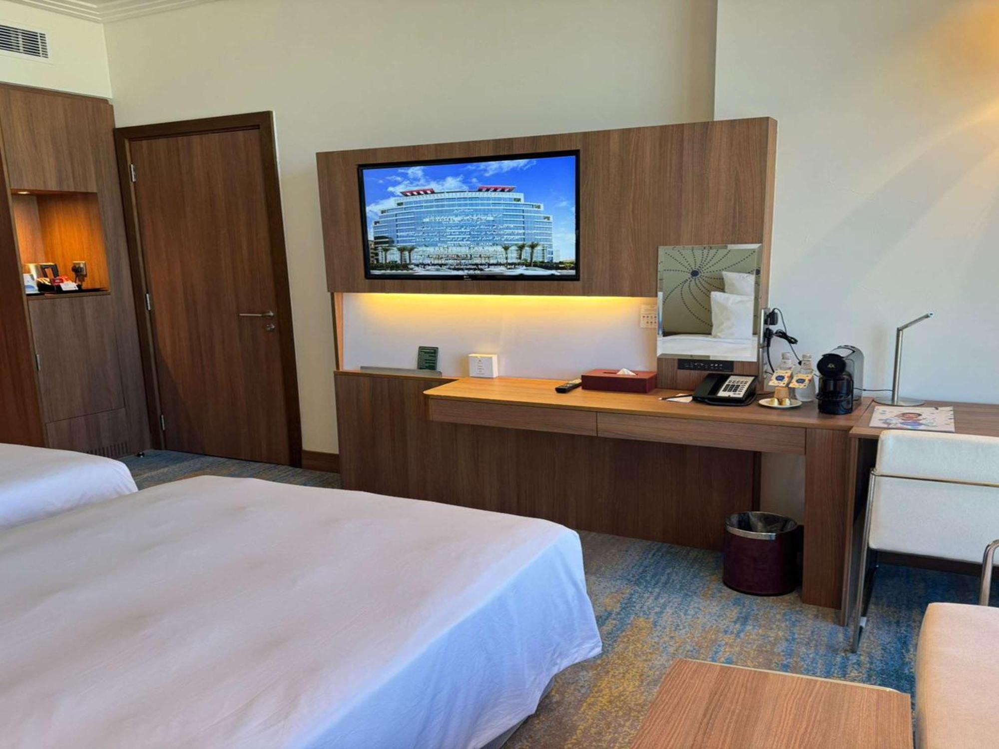 Novotel Jazan Exterior foto The photo shows a hotel room with two beds made up with white linens. There is a wooden wall unit that features a flat-screen television mounted on it, displaying a picture of a large building. Below the TV, there is a desk area with various items, i